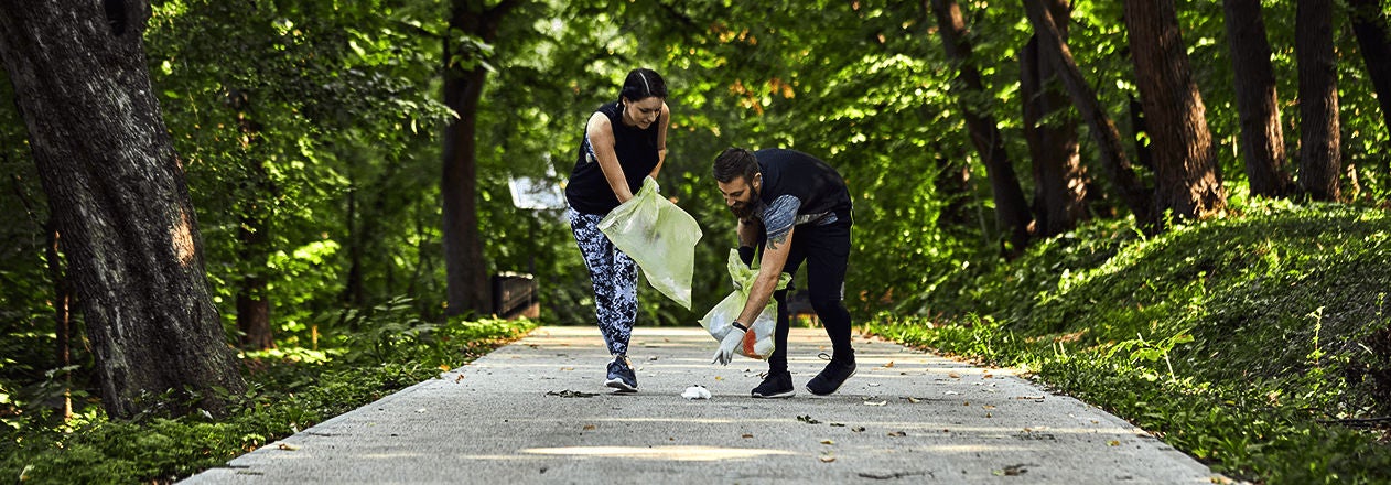 Deux sportifs ramassent des déchets sur un sentier forestier tout en faisant du jogging, illustrant le plogging, une activité qui combine sport et protection de l’environnement 
