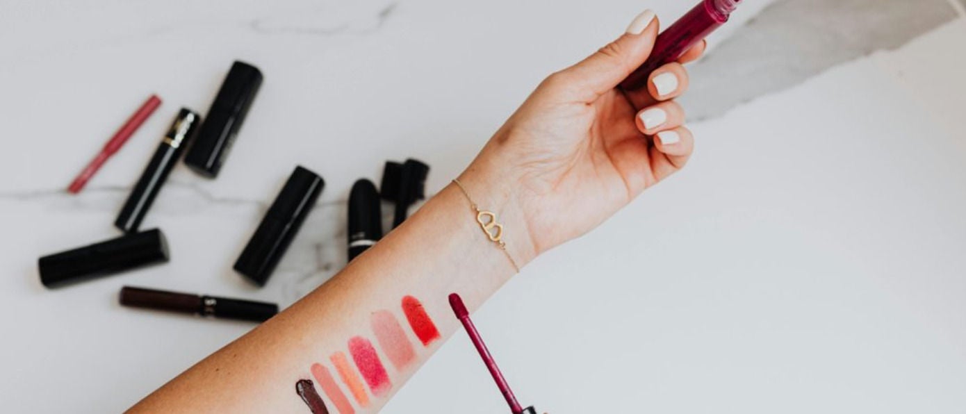 Une personne teste plusieurs couleurs de rouge à lèvres liquide sur son avant-bras, entourée de tubes et crayons de maquillage, illustrant le choix et l’essai de cosmétiques
